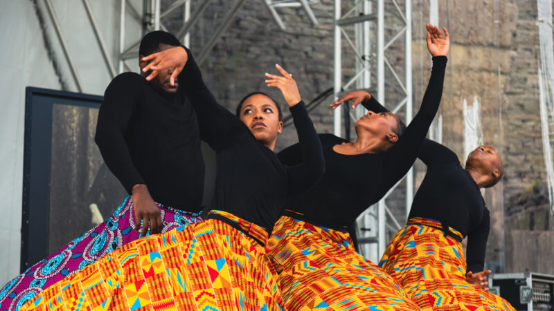 Four Black performers on an outdoor stage wearing black longsleeved tops and full skirts printed with colourful patterns. Their arms are gracefully held in the air and their heads are tilted backwards.