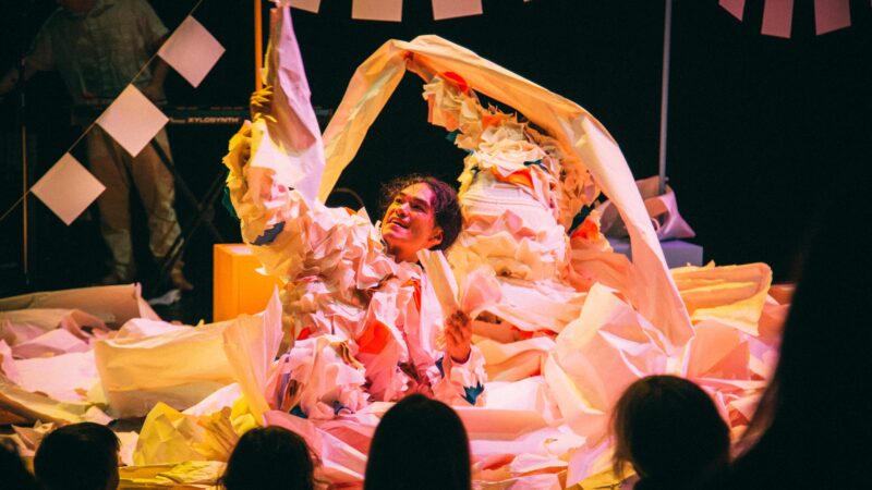 An East Asian performer with long, dark hair tied back lies amongst a layer of folded and crumpled white paper. He is wearing a suit made of crumpled white paper, with flashes of blue and red. He is holding a creased piece of paper high in his left hand, looking towards it with joy. In the background hangs bunting made from pieces of white square shaped pieces of paper. Another performer wearing the same costume is rumaging around in the pile of paper. The lighting is warm and the silhouettes of three audience members' heads are visible in the foreground.