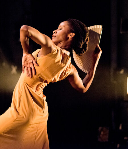 A Black flamenco dancer faces to the left with their black hair tied in a bun and wearing a beige slip dress dances on stage with a beige fan. Their arms are bent at dramatic angles and they are leaning back. The background is black, apart from a warm spotlight that shines horizontally onto the dancer.