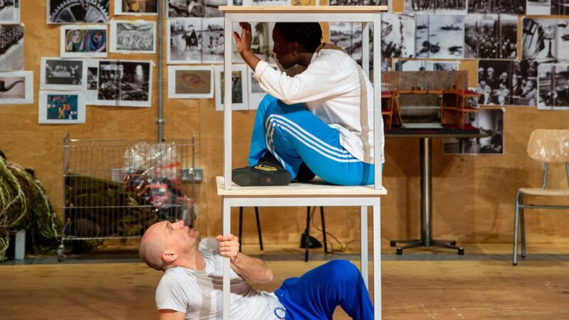 Two square tables stand stacked on top of each other. Underneath the bottom table is a bald, white performer lying on his back and looking up at a Black performer sitting cross legged in the gap underneath the top table. They are both wearing white tops and blue tracksuit bottoms. Behind them is a light wooden wall covered with photographs.