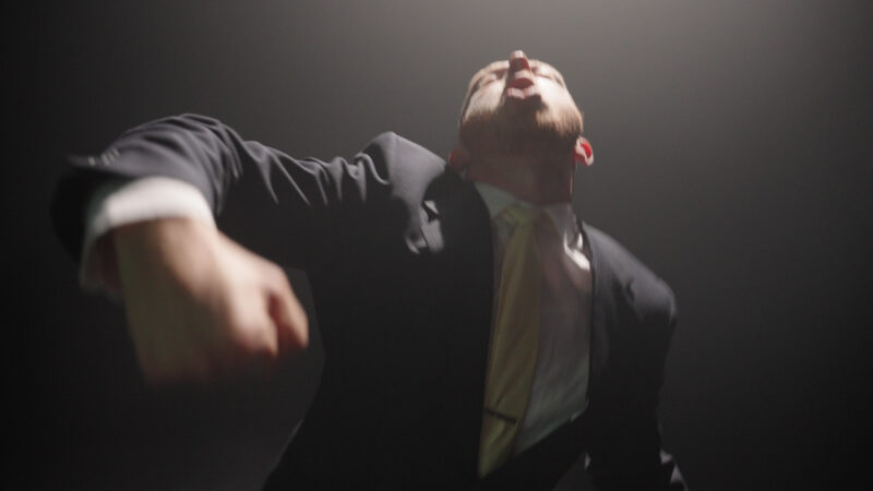 A young white man visible from the waist up is dressed in a black suit, white shirt, and pale yellow tie. He is in motion, flinging his left arm upwards, bent at the elbow with his hand in a fist heavy, pointing downwards. His head is tippe dback, eyes closed, mouth open. The space is dark except the light shining on him and casting shadows on his face.