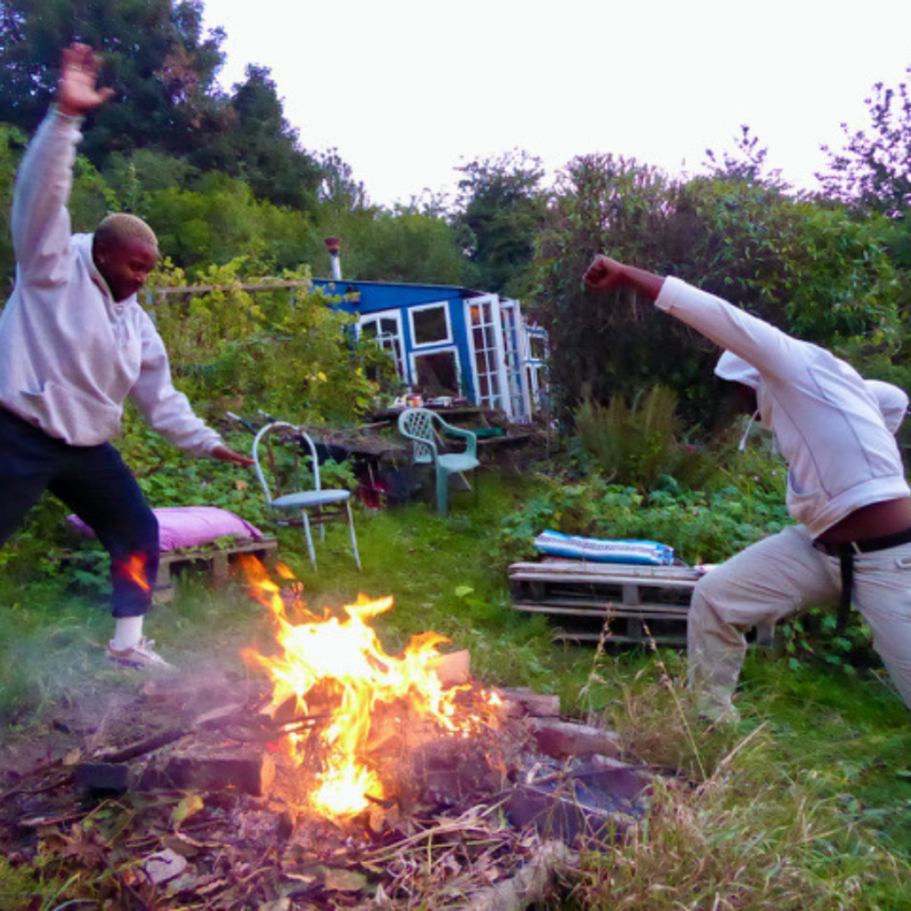 Bakani and Shaq wearing comfortable light clothing making energetic shapes while standing next to a bonfire at dusk. There is a blue garden building in the background surrounded by foliage.