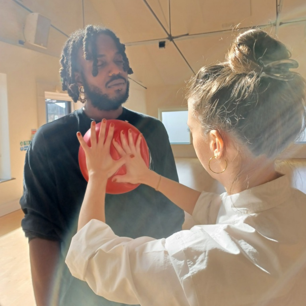 Shaq George being massaged in the centre of his chest by a half inflated medium sized ball held by Jessica Ashley. The ball is red and so is her Jessica's polish. Both figures are in bright sunlight in a dance studio.
