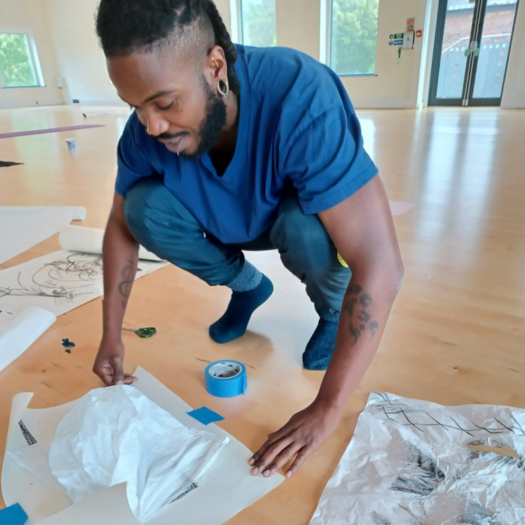 Shaq George in a squat on the floor, using tape to fix crumpled white paper with line drawings and marks to a light wooden floor in an exercise looking into movement scores for improvisation.
