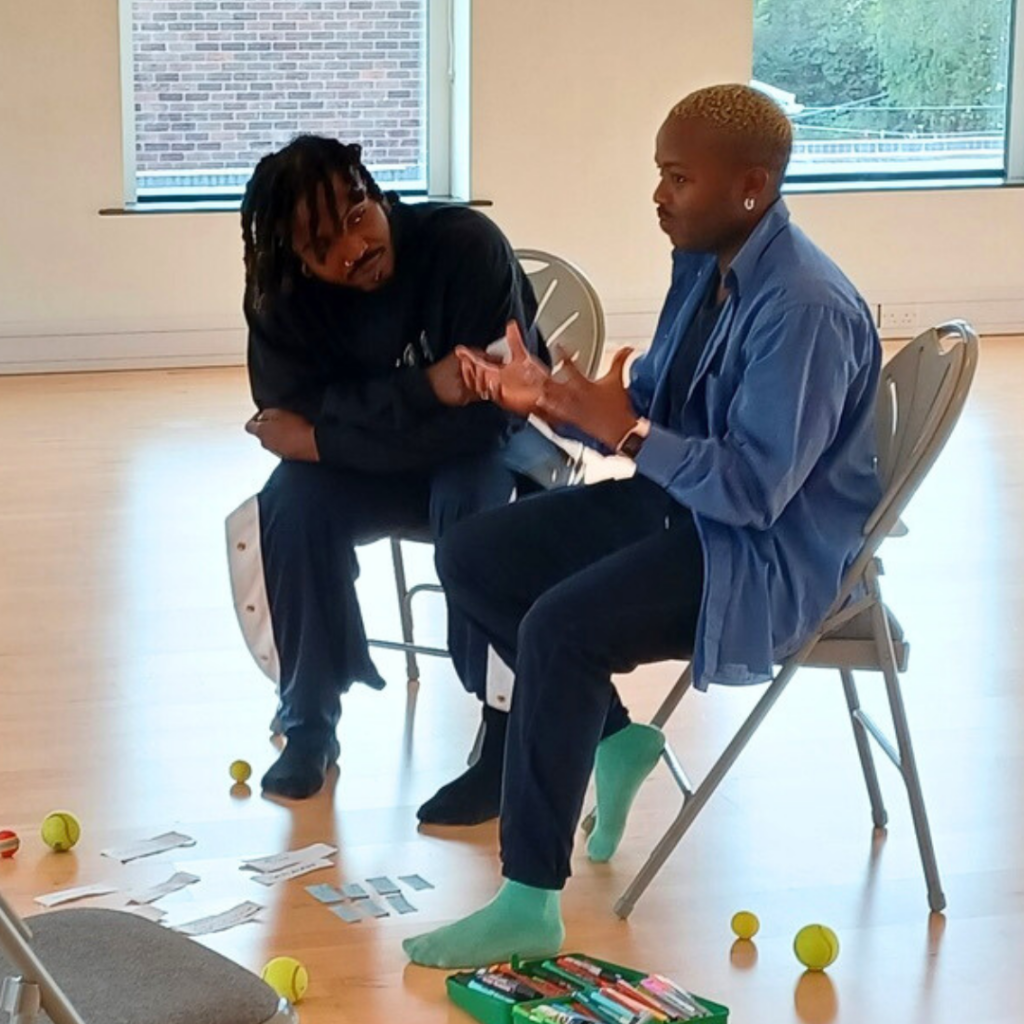 Shaq George and Bakani Pick-Up sat close together on chairs in a dance studio. Shaq is listening intently and Bakani is speaking, using his hands out in front to emphasise his point. There are scraps of paper, pens, and balls scattered around the floor.