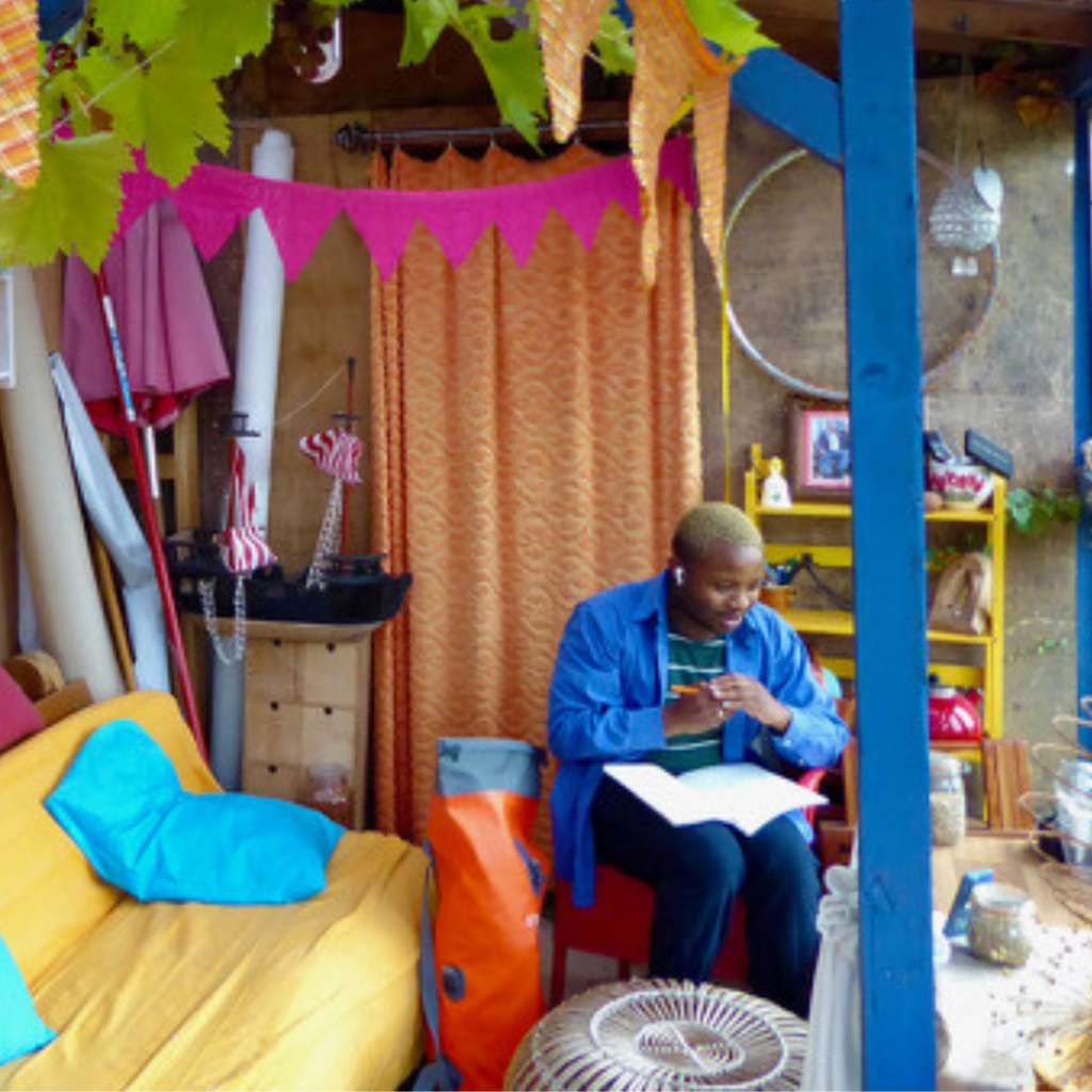 Bakani Pick-Up taking time to reflect alone inside a garden structure made of timber with colourful fabric and pink triangular bunting hung in the background.