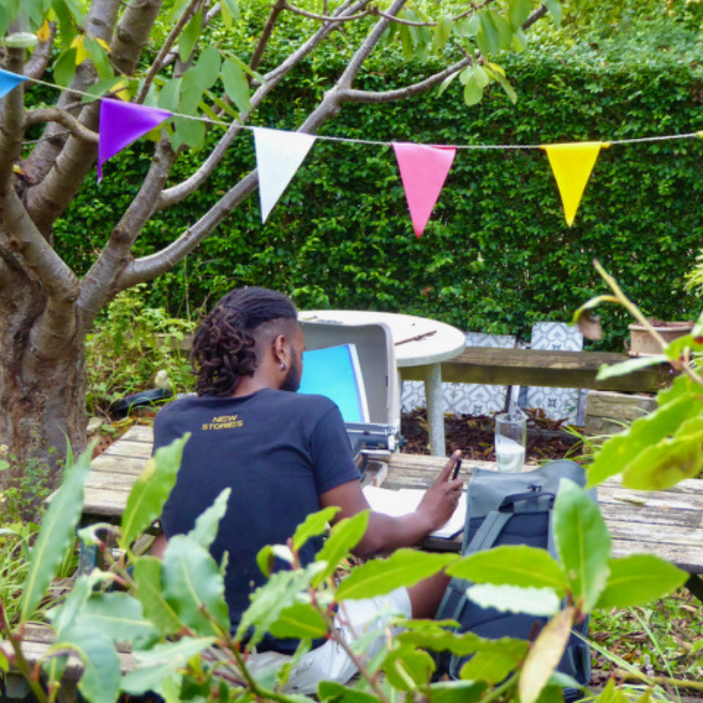 Shaq George sat at a garden bench under a tree with colourful triangular bunting hanging from it. Shaq is writing on paper.