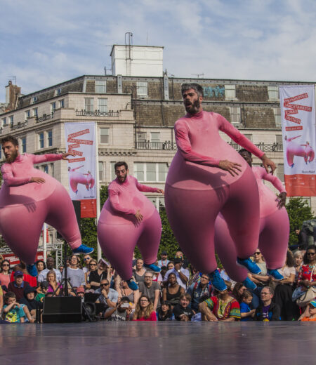 Four men in pink inflatable suits jumping in the air on a stage in front of BIDF flags