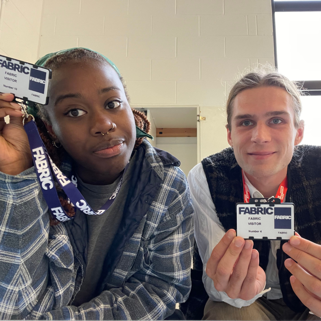 An image of Birmingham CROWD residency artists Chelsea and Hubert holding up their navy FABRIC lanyards and key cards to the camera. Chelsea is wearing a grey and white checked jacket and a grey top, their hair is styled in long braids tied up. She is also wearing a silver septum ring. Hubert is wearing a white shirt and a black gilet, his hair is light brown and tied up.