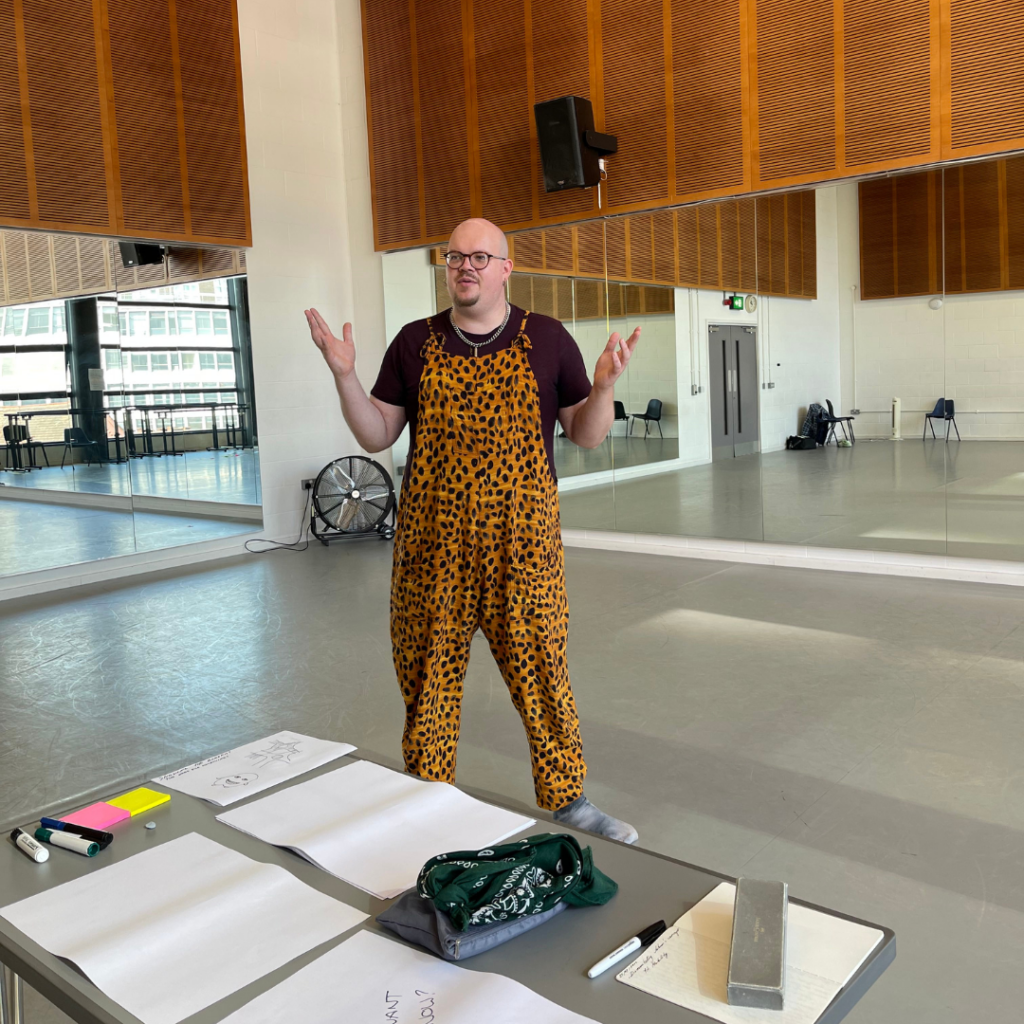 Birmingham based CROWD facilitator Adam Carver stands in a studio with grey floor, mirrored walls, and wood panelling above the mirrors. Adam is wearing a black t-shirt, and orange animal print dungarees. Adam is white, bald, and is wearing glasses. In front of Adam is a table with paper and pens scattered on it.