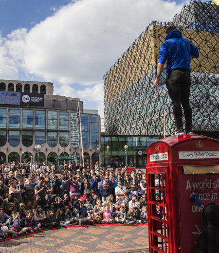 Man in blue top standing on top of a bright red phone box in front of a crowd of viewers in front of the Library of Birmingham