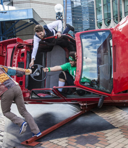 Red car on its side with three people hanging from it by holding onto the steering wheel