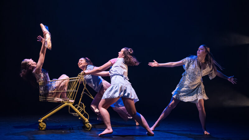 One dancer sitting in a bright yellow shopping trolley holding a baguette in the air. Three other dancers pulling the trolley towards them.