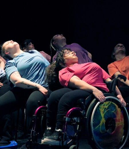 Dancers in colourful t-shirts, some in wheelchairs, some sitting on a chair, some squatting. They all lean to the left looking upwards.