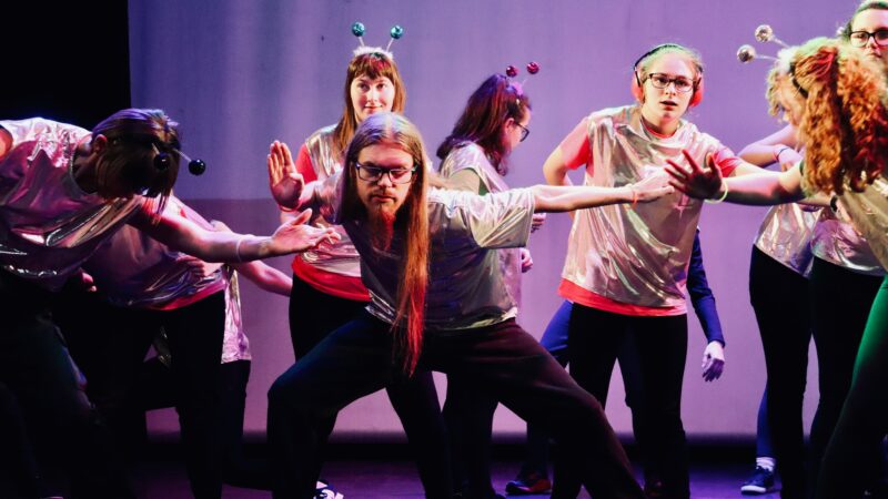 A mixed gender group of dancers performing on a purple lit stage