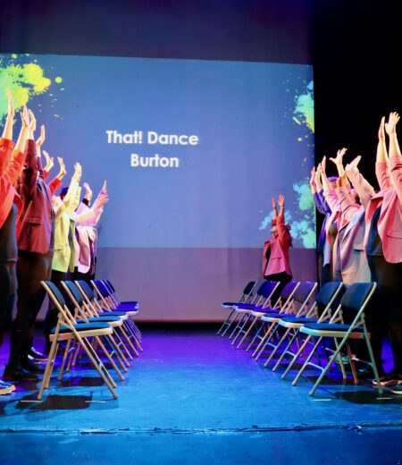 Two lines of dancers on a colourful lit stage standing face to face behind chairs with their arms up.