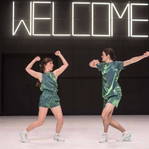 A dance partnership on stage mid-performance dancing below a neon sign that reads