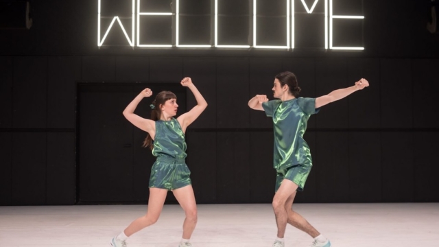 A dance partnership on stage mid-performance dancing below a neon sign that reads "Welcome".
