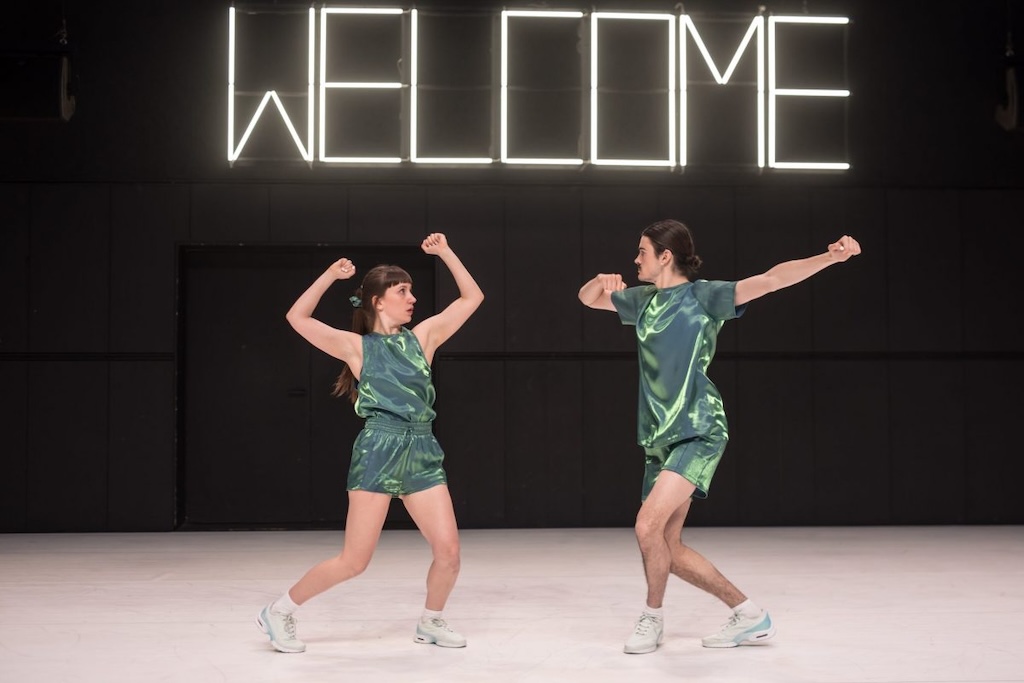 A dance partnership on stage mid-performance dancing below a neon sign that reads "Welcome".