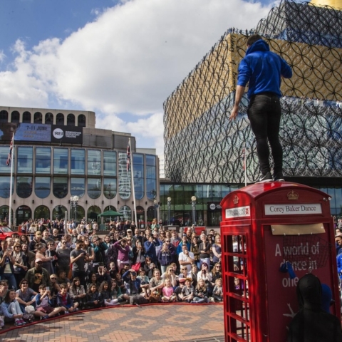 A person stood on a red telephone box with a crowd watching them.