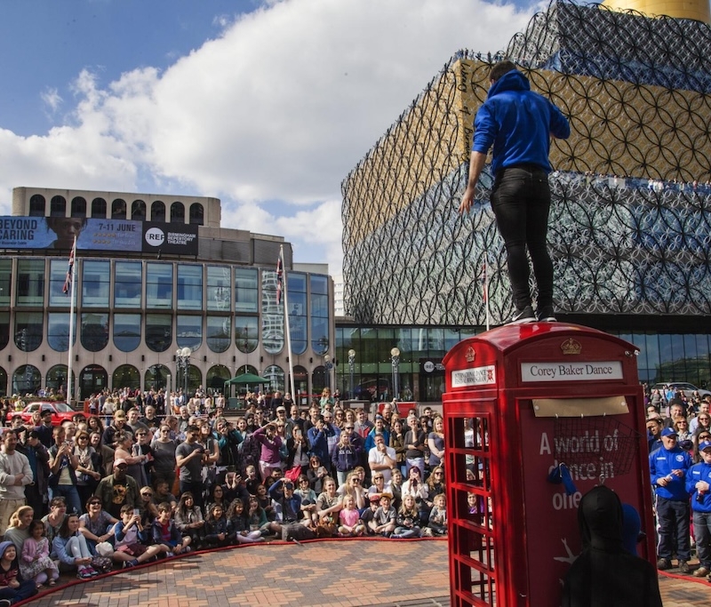 A person stood on a red telephone box with a crowd watching them.