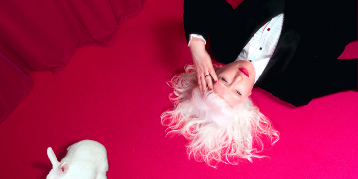 A photograph of Jo Bannon lying on a vibrant pink carpet with her white hair fanned out around her head. She is wearing a black suit and white shirt and hot pink lipstick. Her hand is held to her forehead. A small white rabbit is positioned nearby. The background features a rich pink curtain.