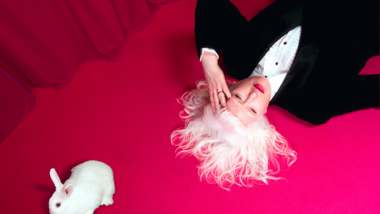 A photograph of Jo Bannon lying on a vibrant pink carpet with her white hair fanned out around her head. She is wearing a black suit and white shirt and hot pink lipstick. Her hand is held to her forehead. A small white rabbit is positioned nearby. The background features a rich pink curtain.
