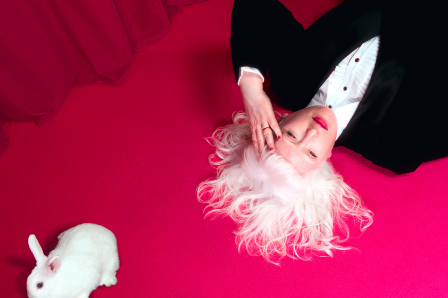 A photograph of Jo Bannon lying on a vibrant pink carpet with her white hair fanned out around her head. She is wearing a black suit and white shirt and hot pink lipstick. Her hand is held to her forehead. A small white rabbit is positioned nearby. The background features a rich pink curtain.