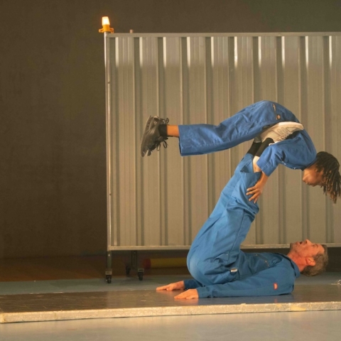 A dance duo mid-performance. One performer is laid on their back with their legs in the air holding the other person up with their feet.